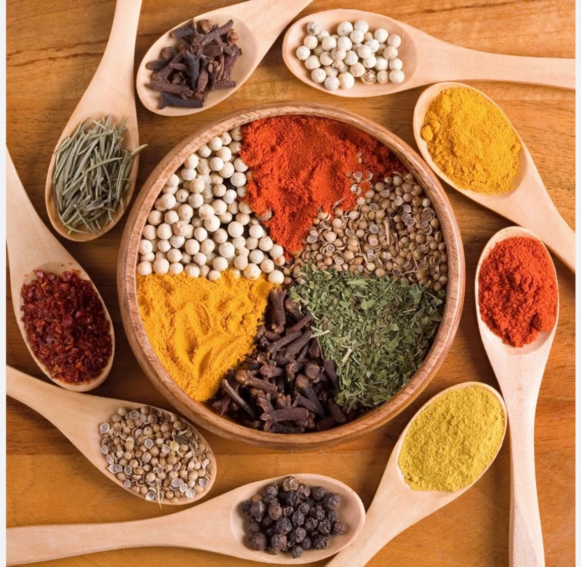 Assorted whole and ground spices in a wooden bowl surrounded by spoons of turmeric, cloves, peppercorns and coriander